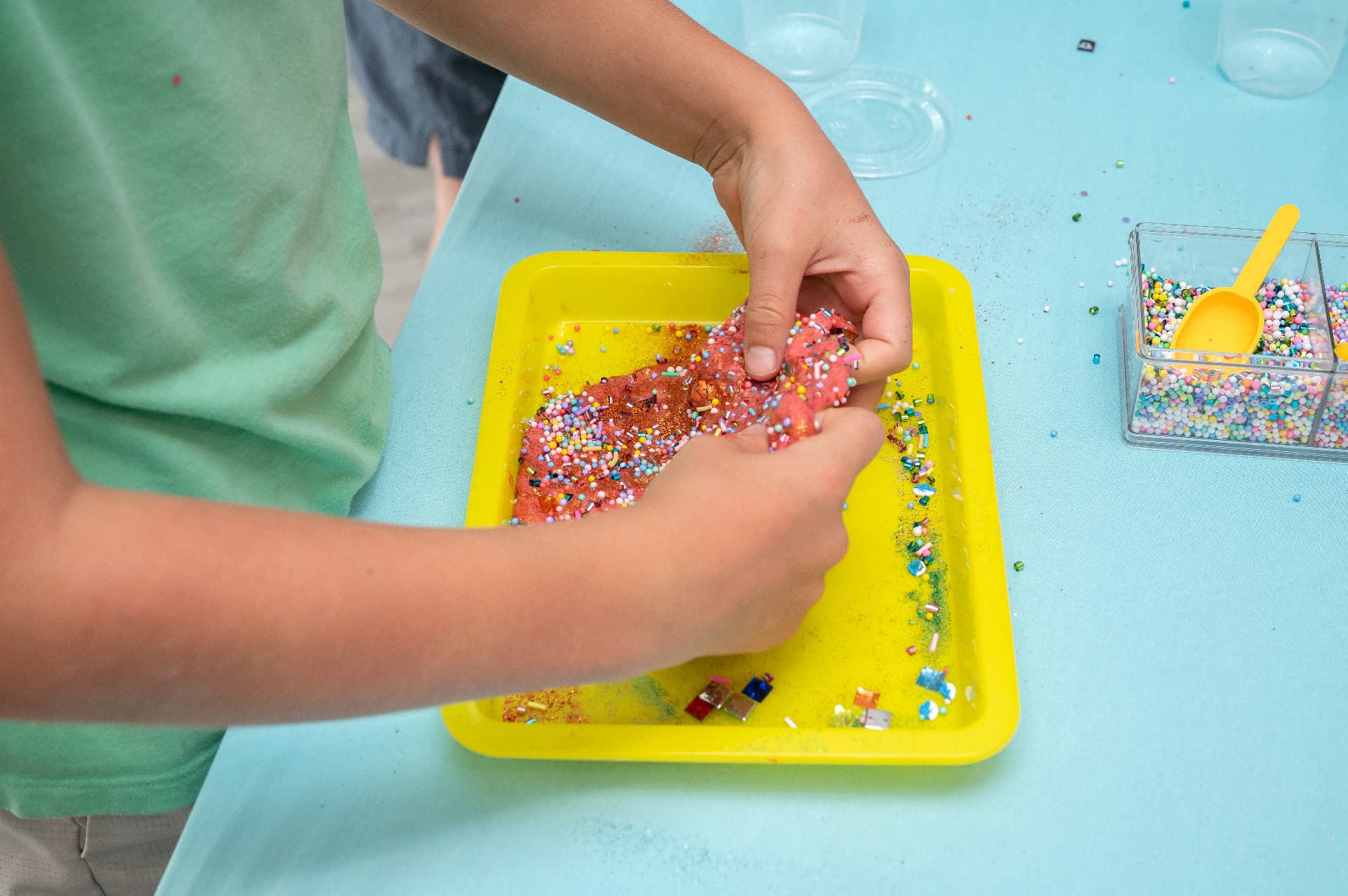Kid mixing glitter and foam beads into slime on a yellow tray