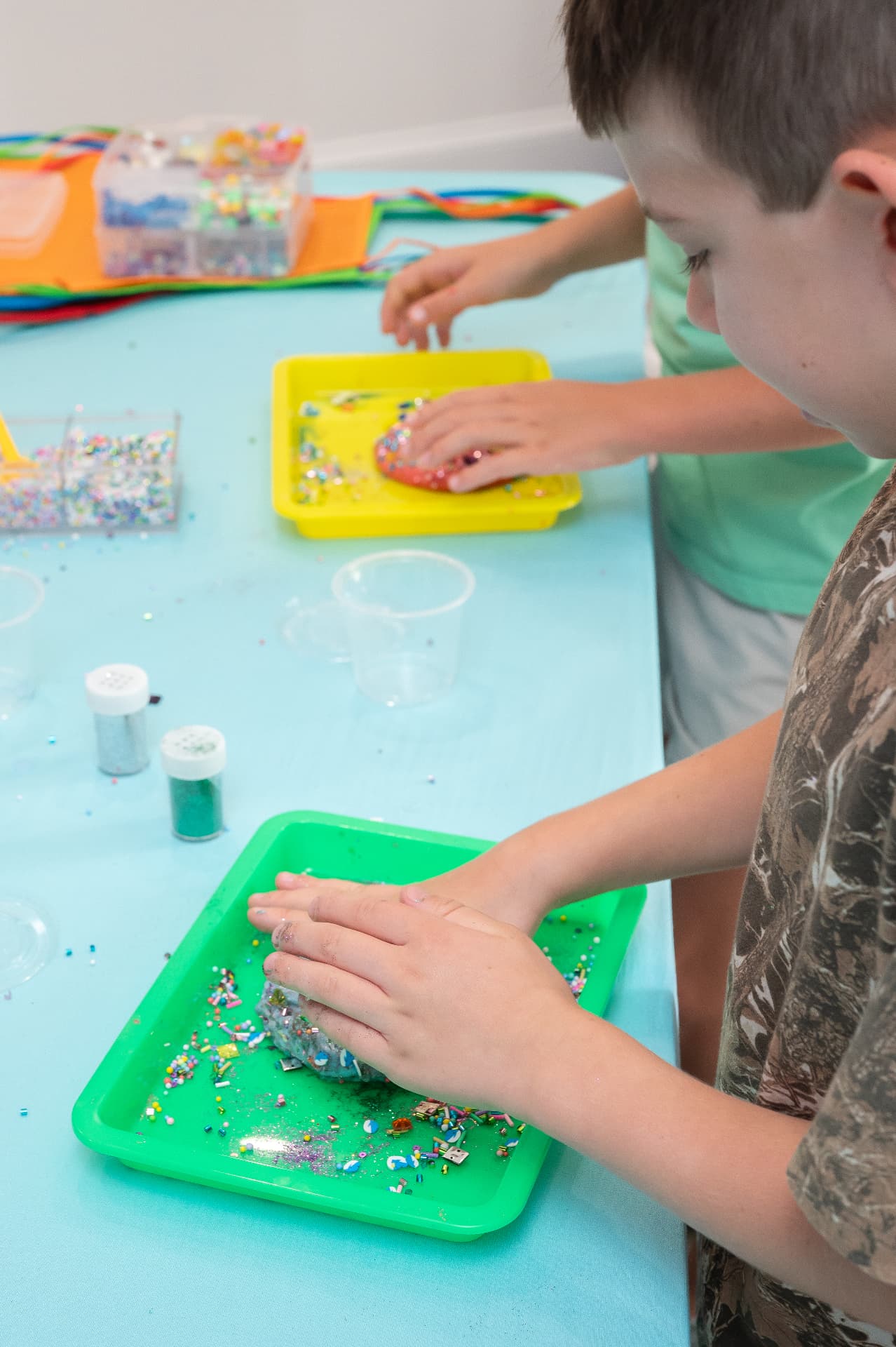 Kids mixing slime on colorful trays with glitter shakers